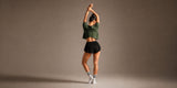 Woman stretching both arms overhead, seen from behind, wearing the KYDRA Cross Back Tee in Forest with Pleated Shorts in Black and striped crew socks, against a warm taupe studio backdrop.