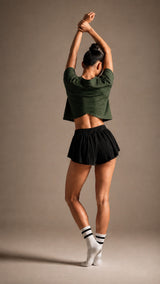 Woman stretching both arms overhead, seen from behind, wearing the KYDRA Cross Back Tee in Forest with Pleated Shorts in Black and striped crew socks, against a warm taupe studio backdrop.